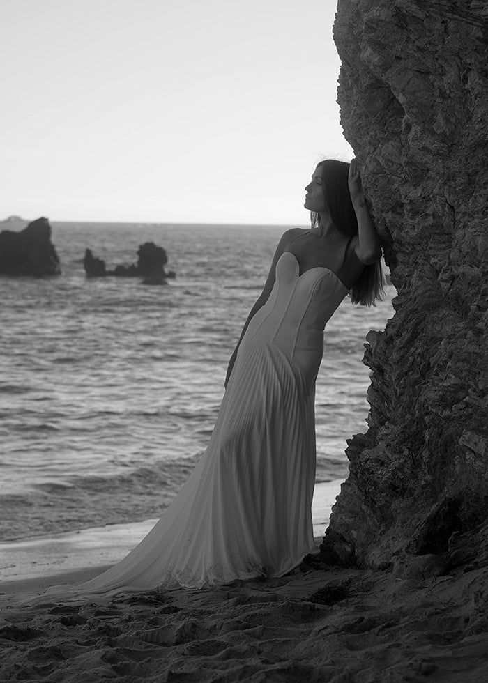 
                  
                    Bride wears crepe corset a-line pleated wedding dress while leaning on a rock on the beach.
                  
                