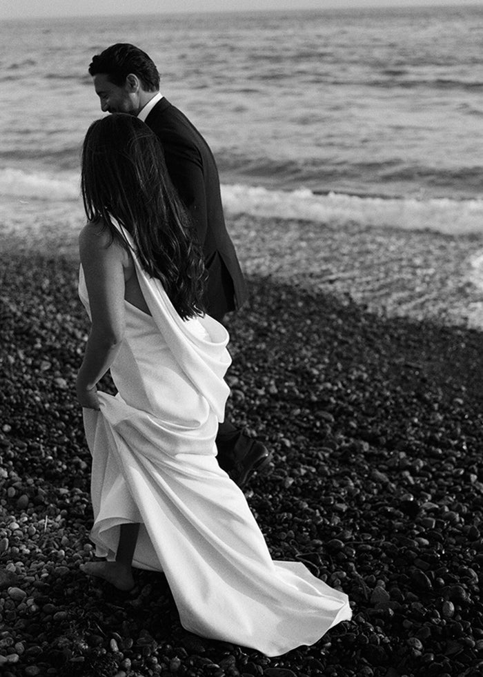 Bride walks barefoot on the beach wearing one shoulder stretch satin wedding dress with cowl back details for summer wedding