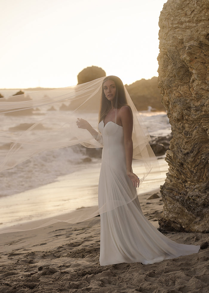 Bride wears custom strapless corset pleated crepe a-line wedding dress while wearing cathedral tulle veil on the beach
